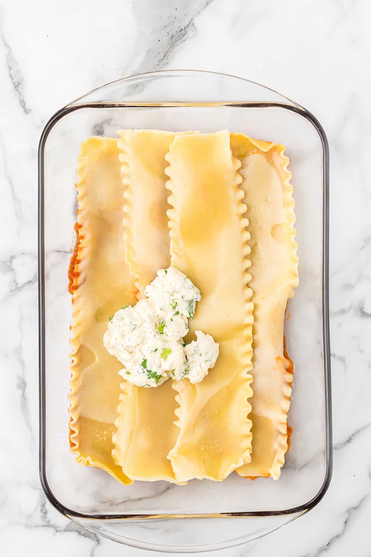 A glass dish with lasagna noodles, tomato sauce, and a dollop of cheese filling on a marble surface.