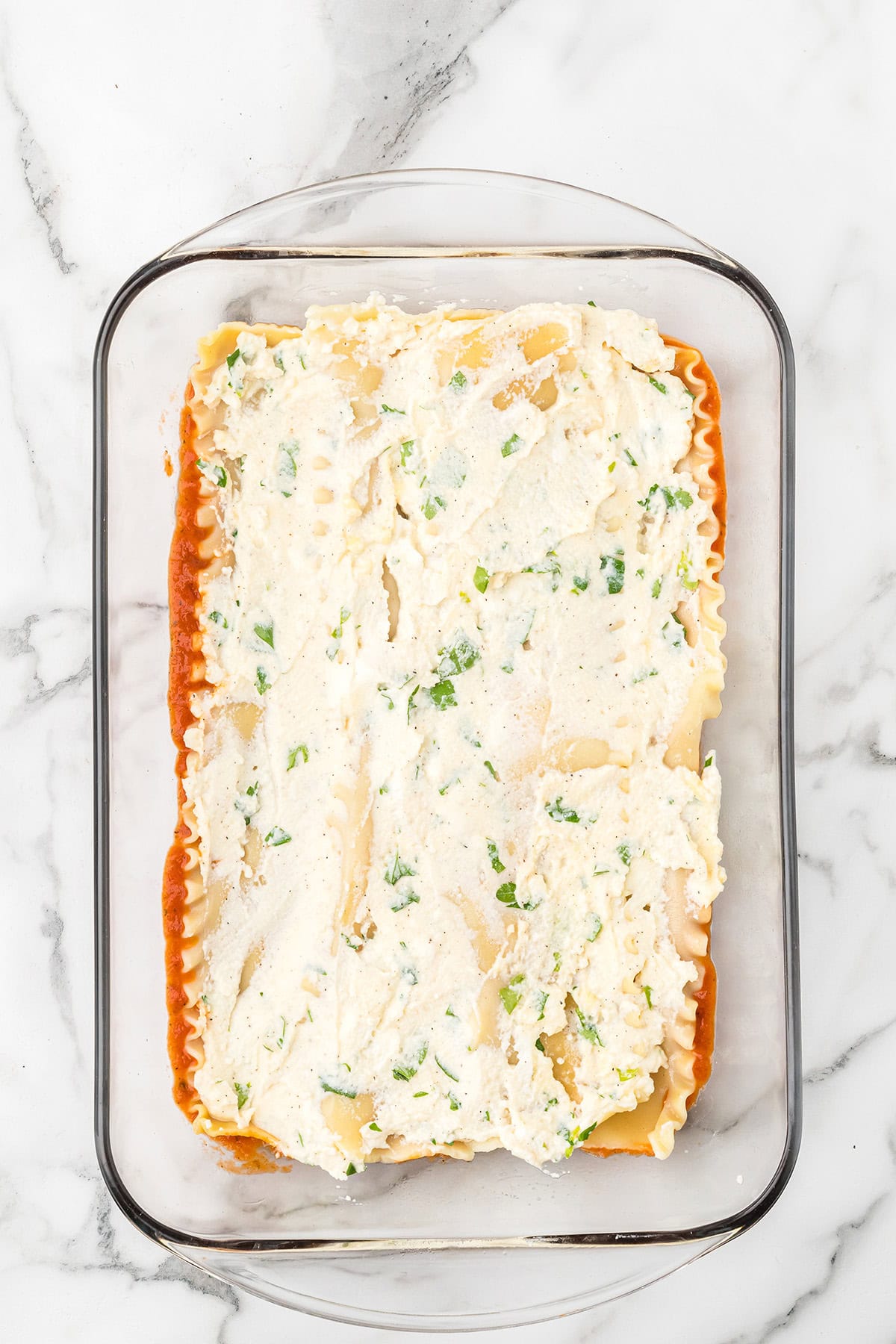Glass baking dish with a layer of lasagna noodles and ricotta cheese mixture on tomato sauce.