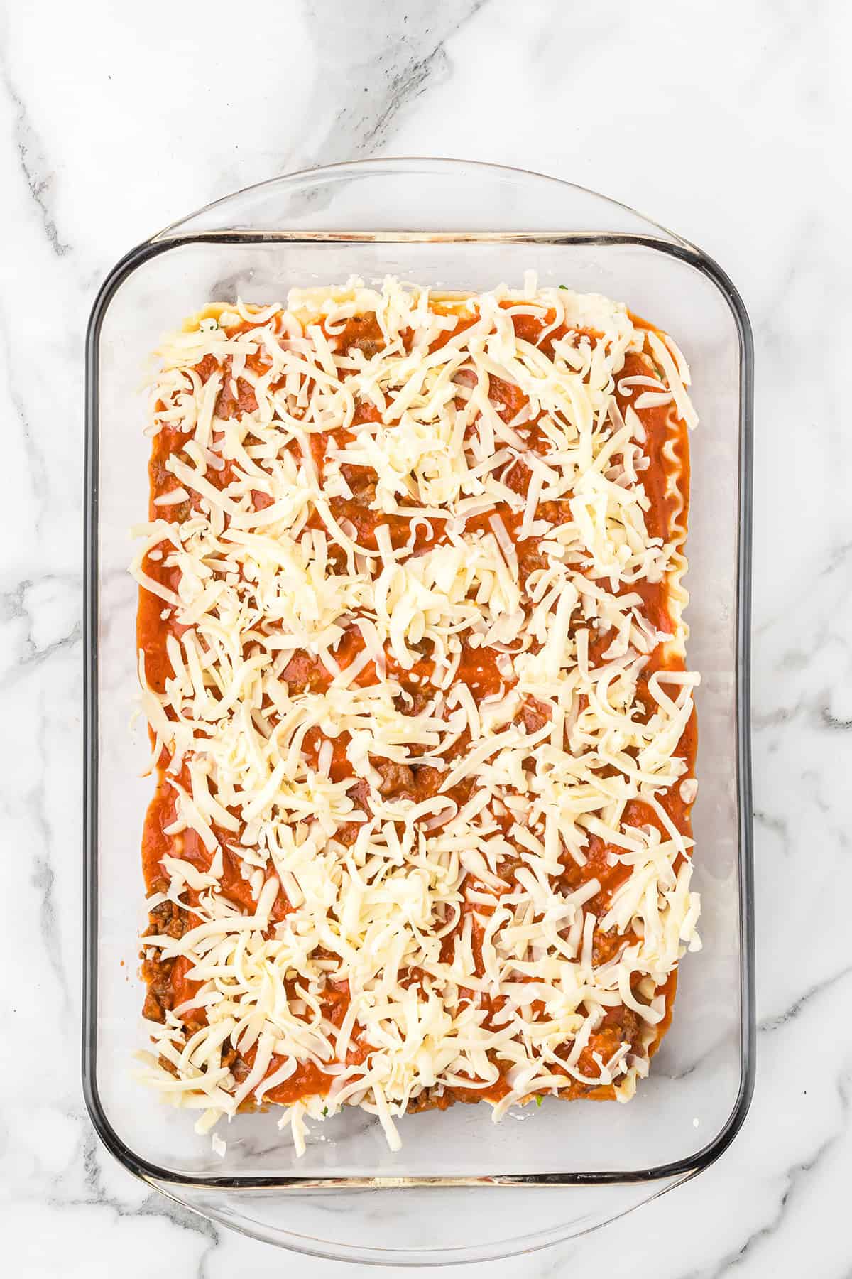 Unbaked lasagna in a glass dish, topped with shredded cheese on a marble surface.
