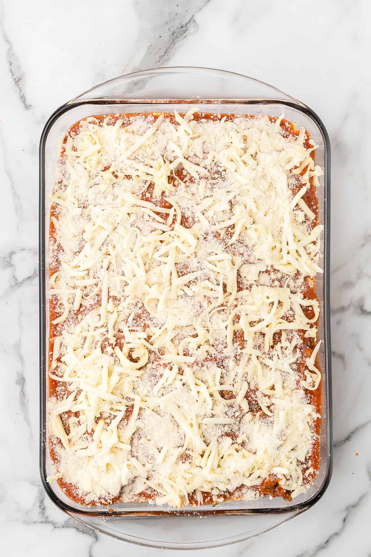 Glass baking dish filled with lasagna topped with shredded cheese and sauce, on a marble countertop.