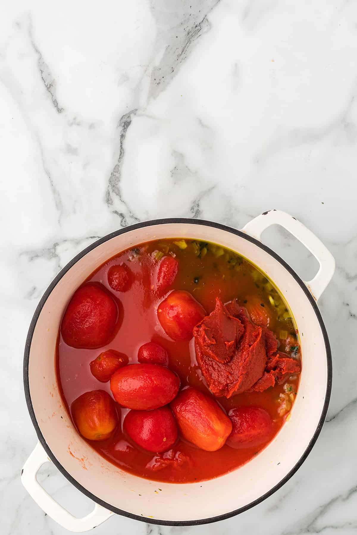 White pot with whole peeled tomatoes and tomato paste on a marble countertop, ready for cooking.