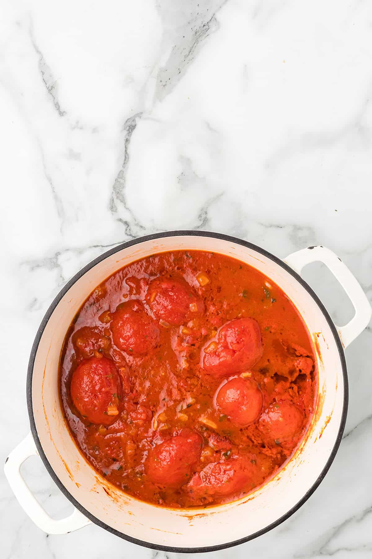 White pot filled with whole peeled tomatoes in red sauce on a marble countertop, viewed from above.