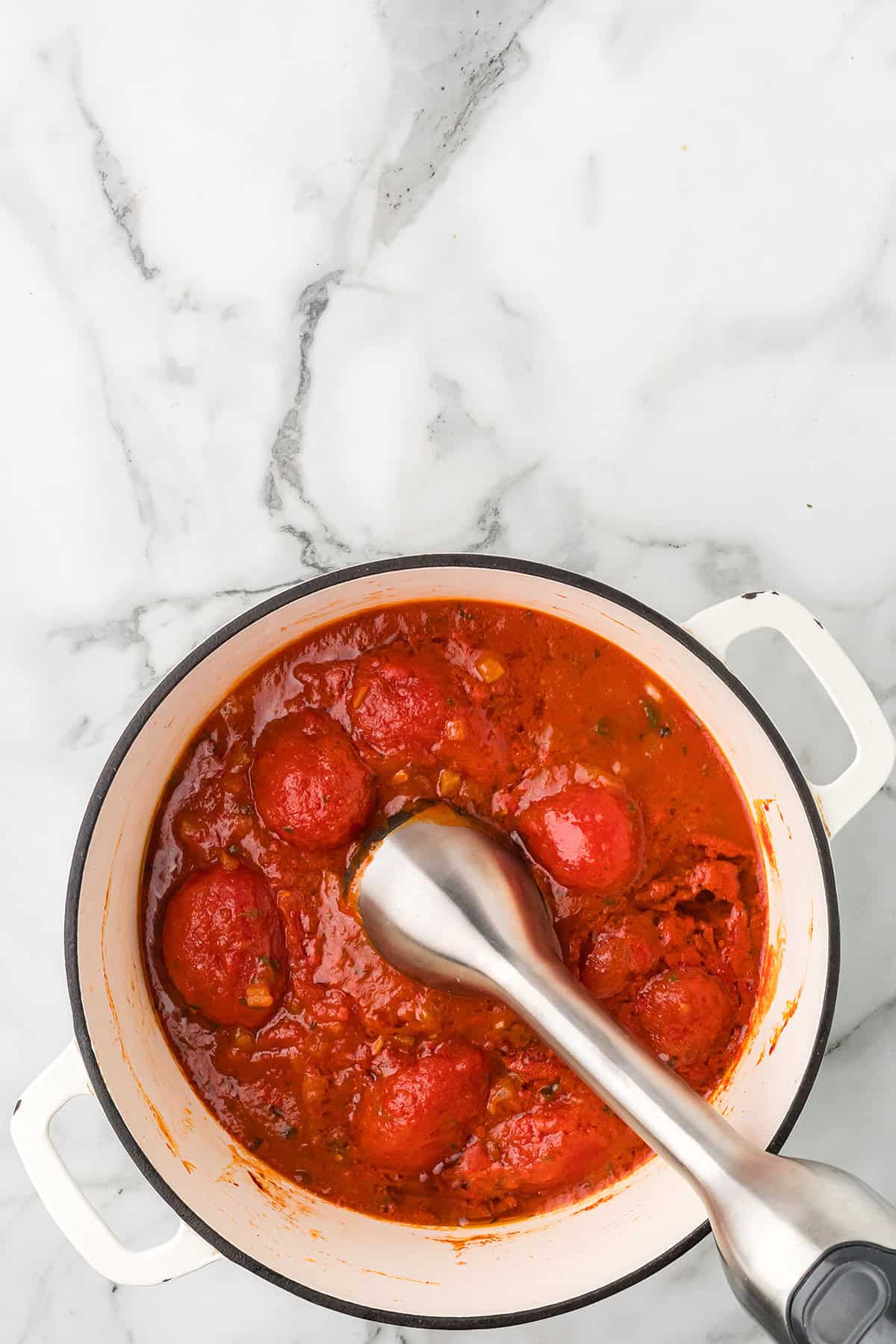 A white pot of tomato sauce with whole tomatoes and an immersion blender on a marble surface.
