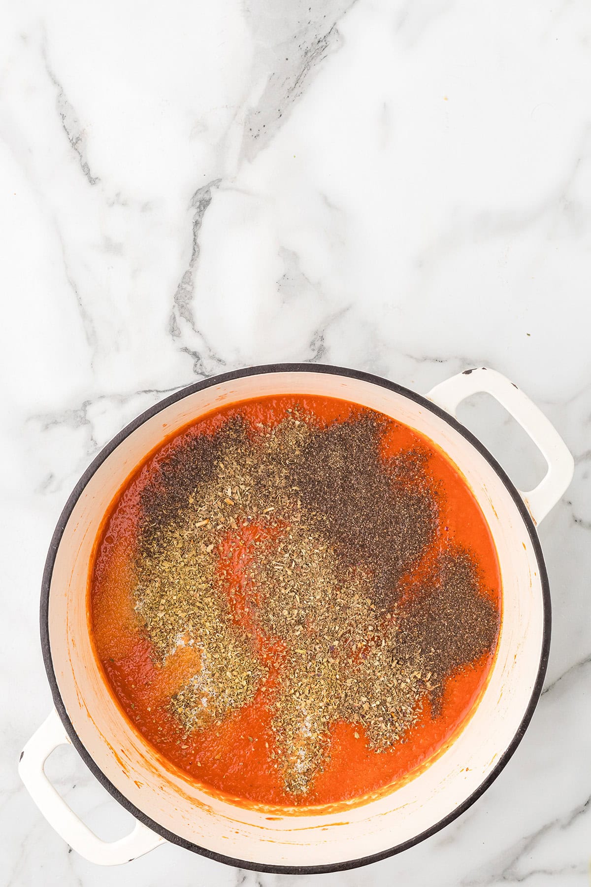 A pot of tomato sauce topped with dried herbs sits on a white marble surface.