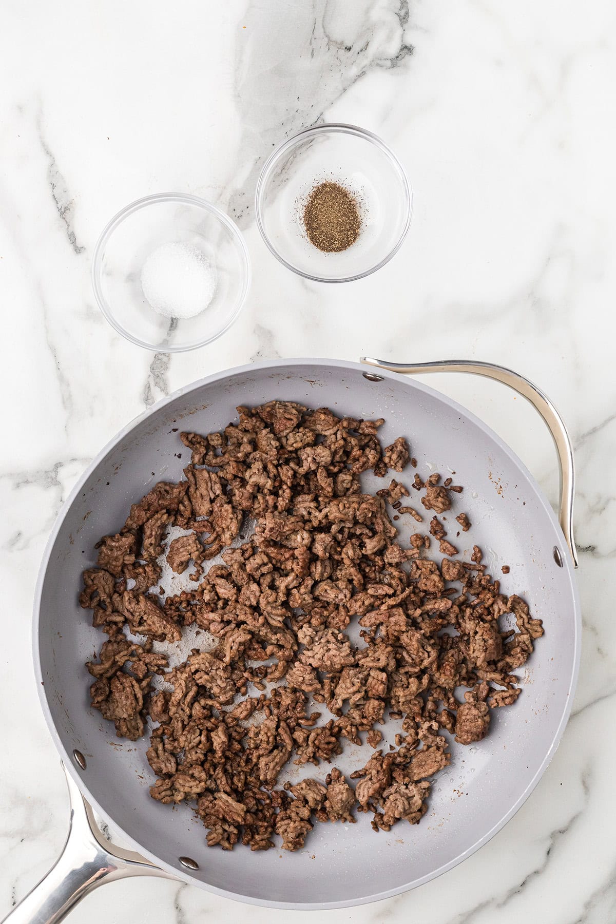 Cooked ground beef in a skillet on a marble counter, with bowls of salt and pepper nearby.