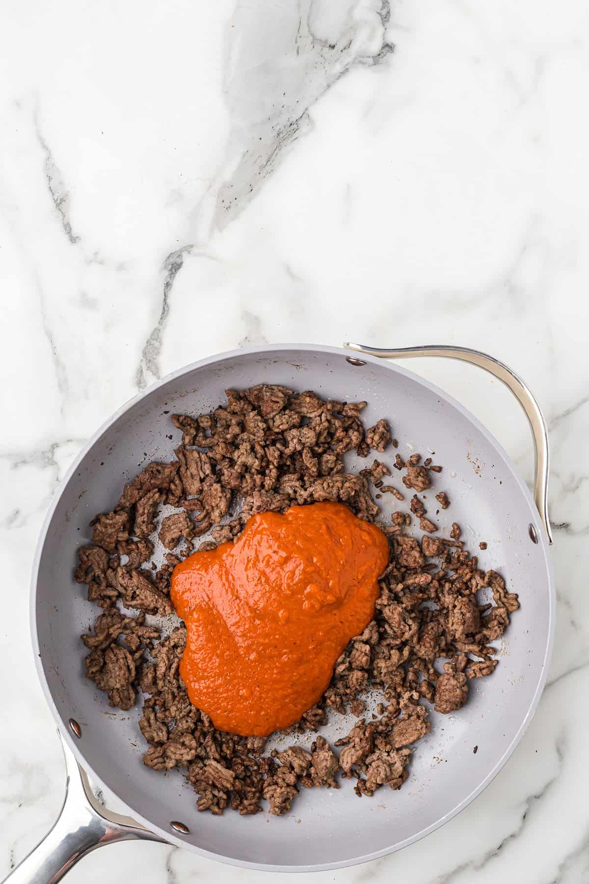 Ground beef in a skillet topped with marinara sauce, on a white marble countertop.