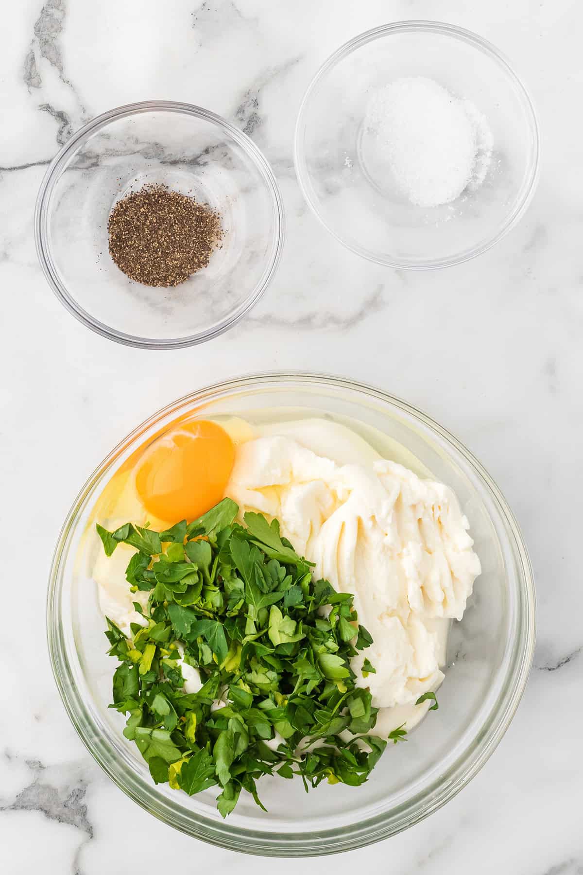 A bowl with ricotta, an egg, and chopped parsley; bowls of salt and pepper beside it on marble.