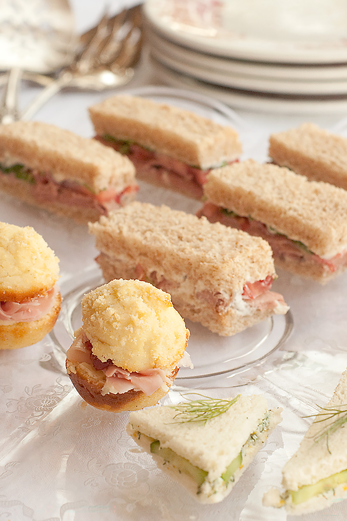 A plate with assorted finger sandwiches and savory pastries is arranged on a white tablecloth with plates and utensils in the background.