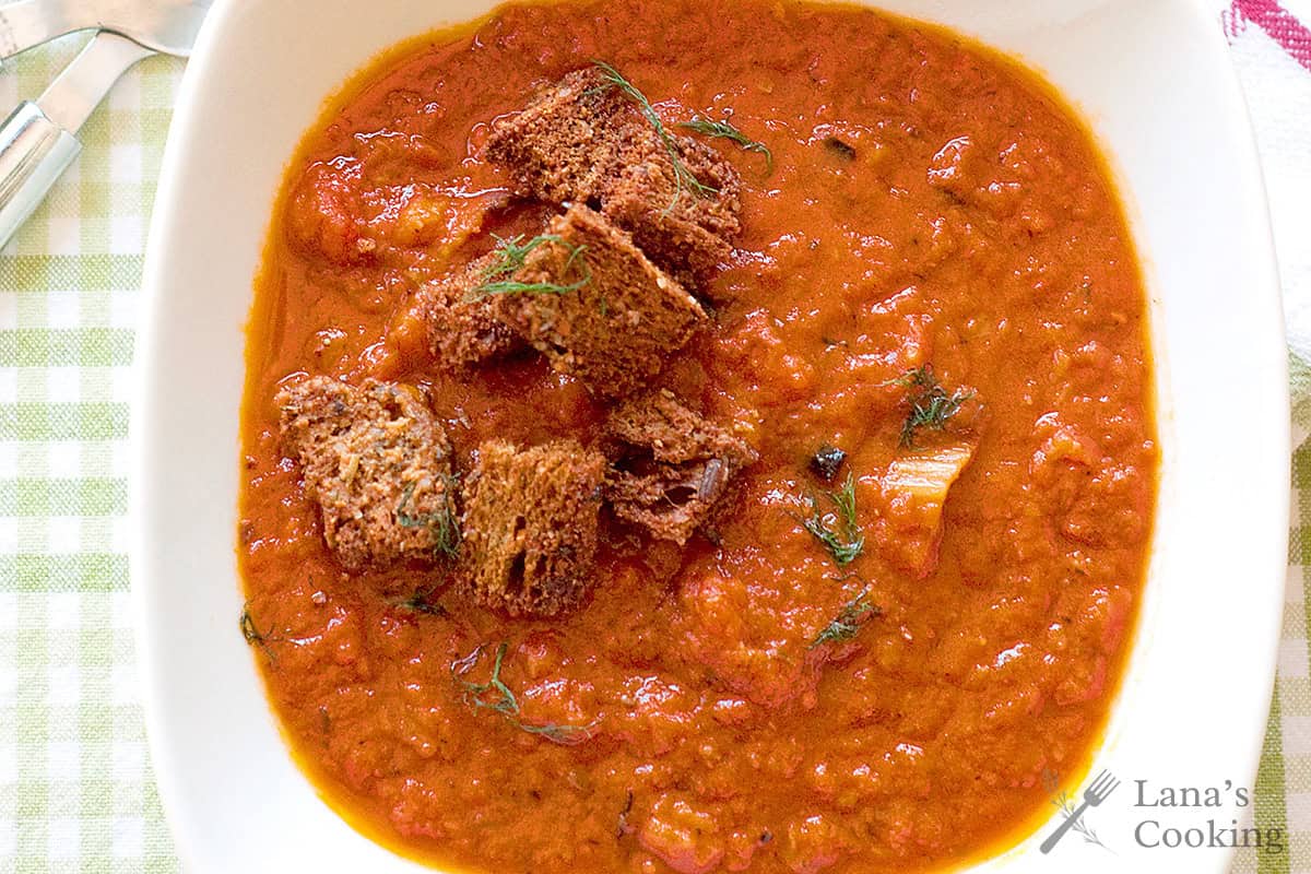 Thick tomato soup topped with croutons and herbs, served in a white bowl on a checkered tablecloth.