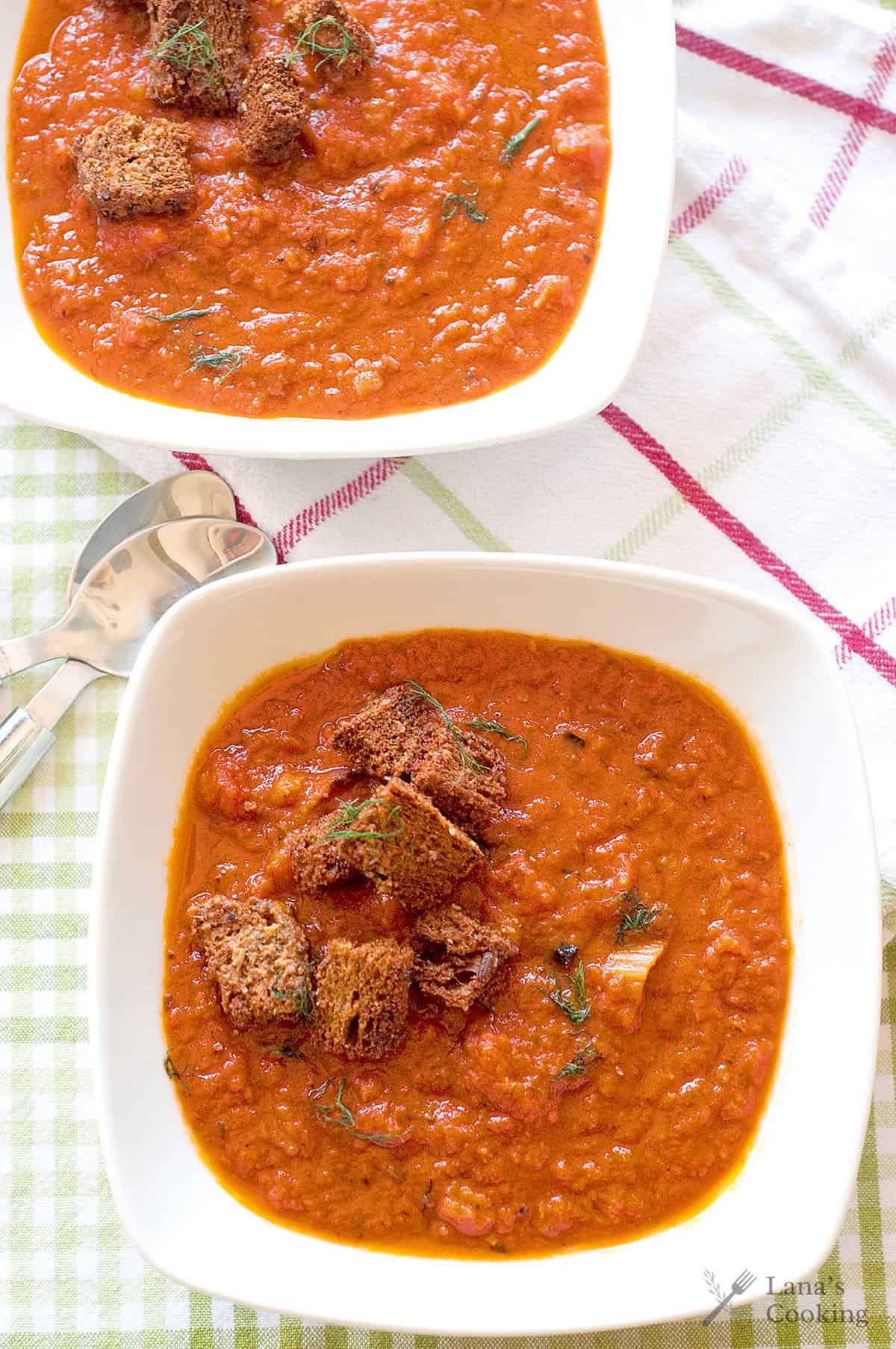 Two bowls of tomato soup topped with croutons and herbs, placed on a checkered cloth with spoons nearby.