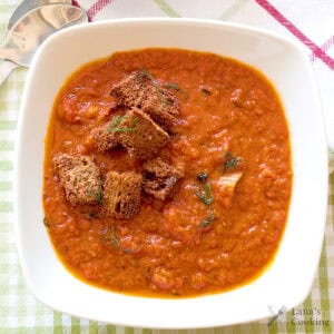 A bowl of tomato fennel soup topped with croutons and herbs, on a checkered tablecloth with a spoon nearby.