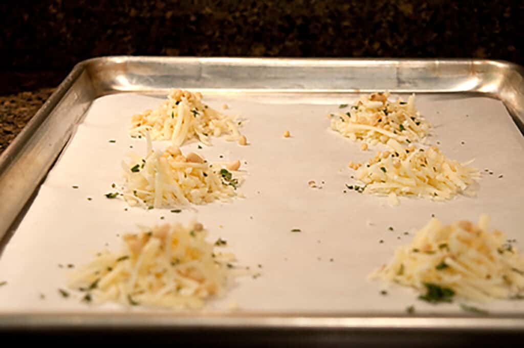 Mounds of grated cheese with sage and nuts on a parchment-lined pan.