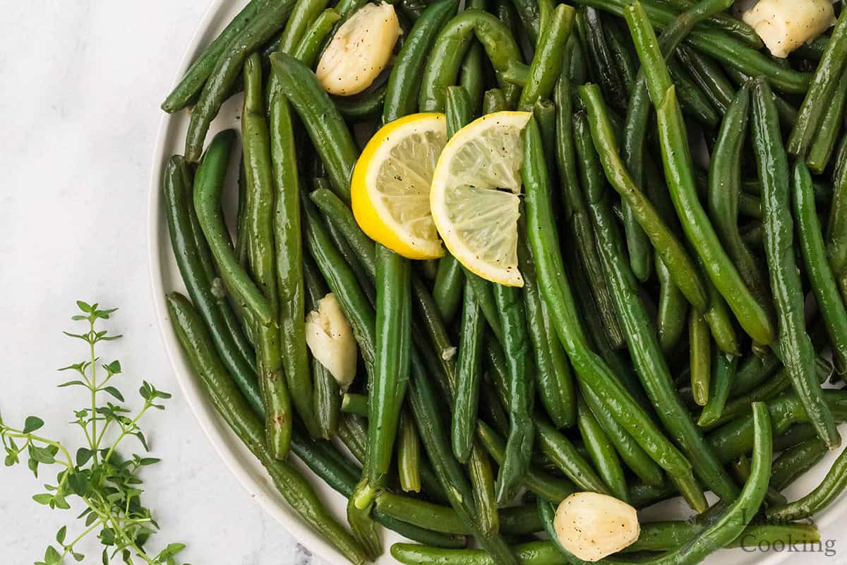 Green beans with garlic cloves and lemon slices on a plate, garnished with fresh thyme on the side.