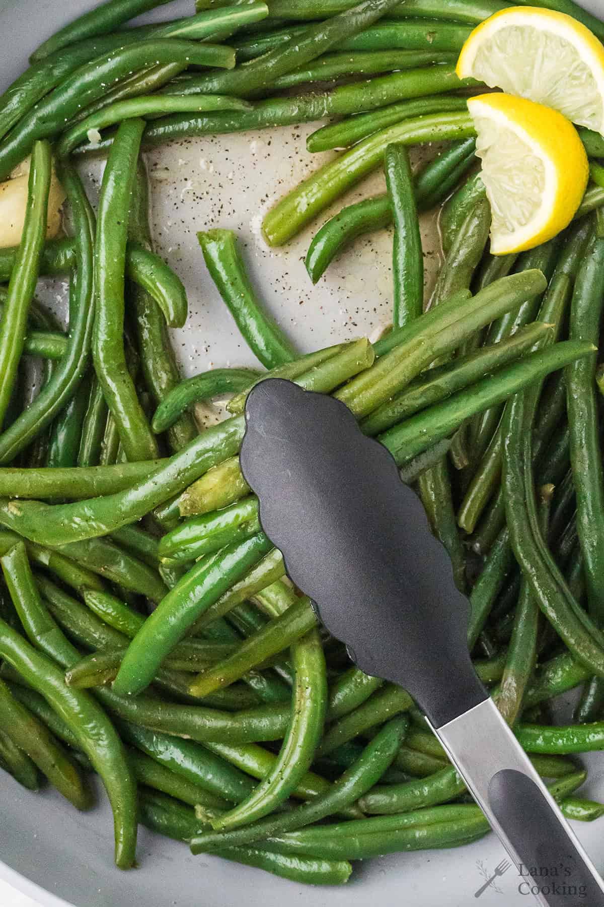 Green beans being sautéed in a pan with black tongs and lemon wedges visible.