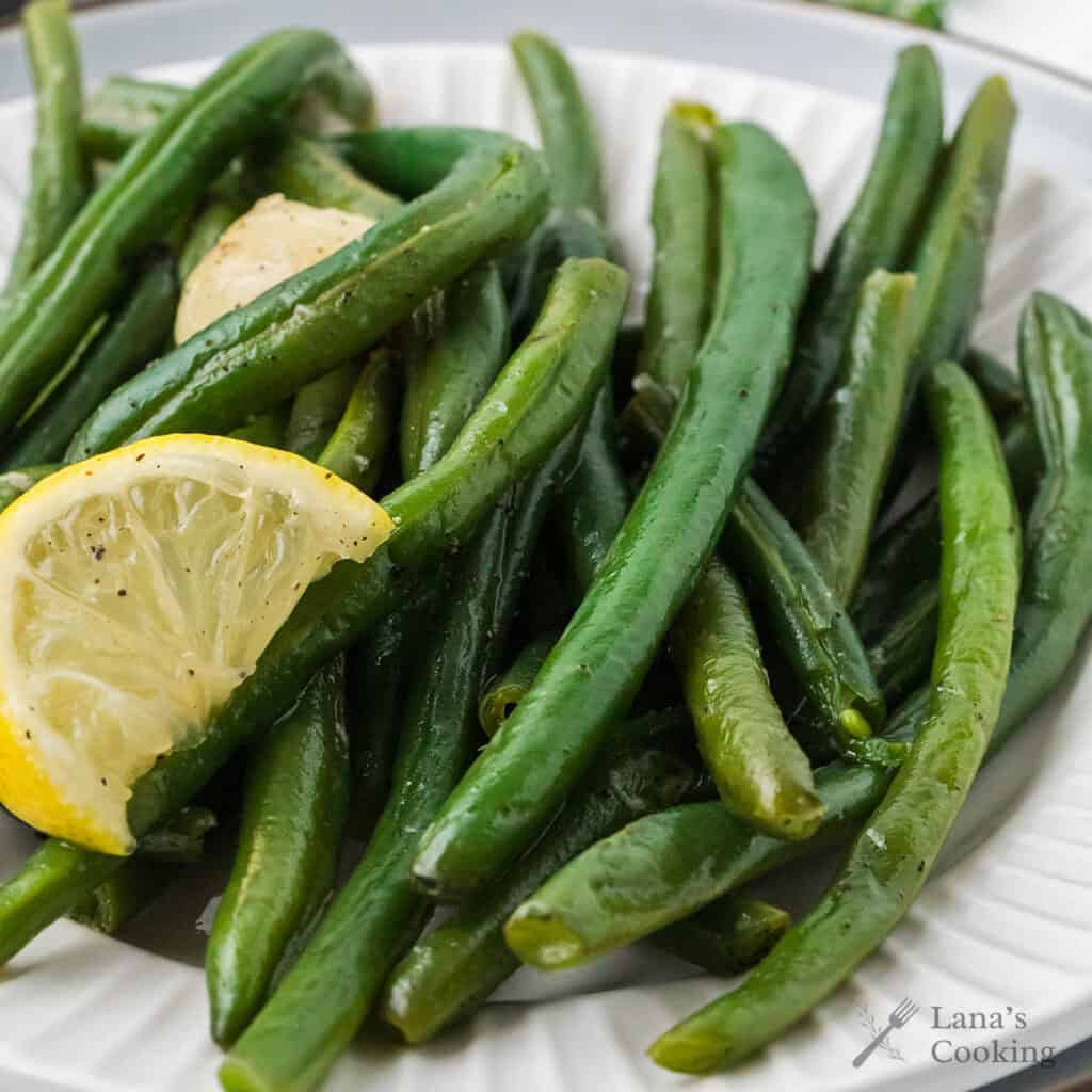 Green beans garnished with a lemon wedge and garlic on a white plate.