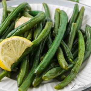 Green beans garnished with a lemon wedge and garlic on a white plate.