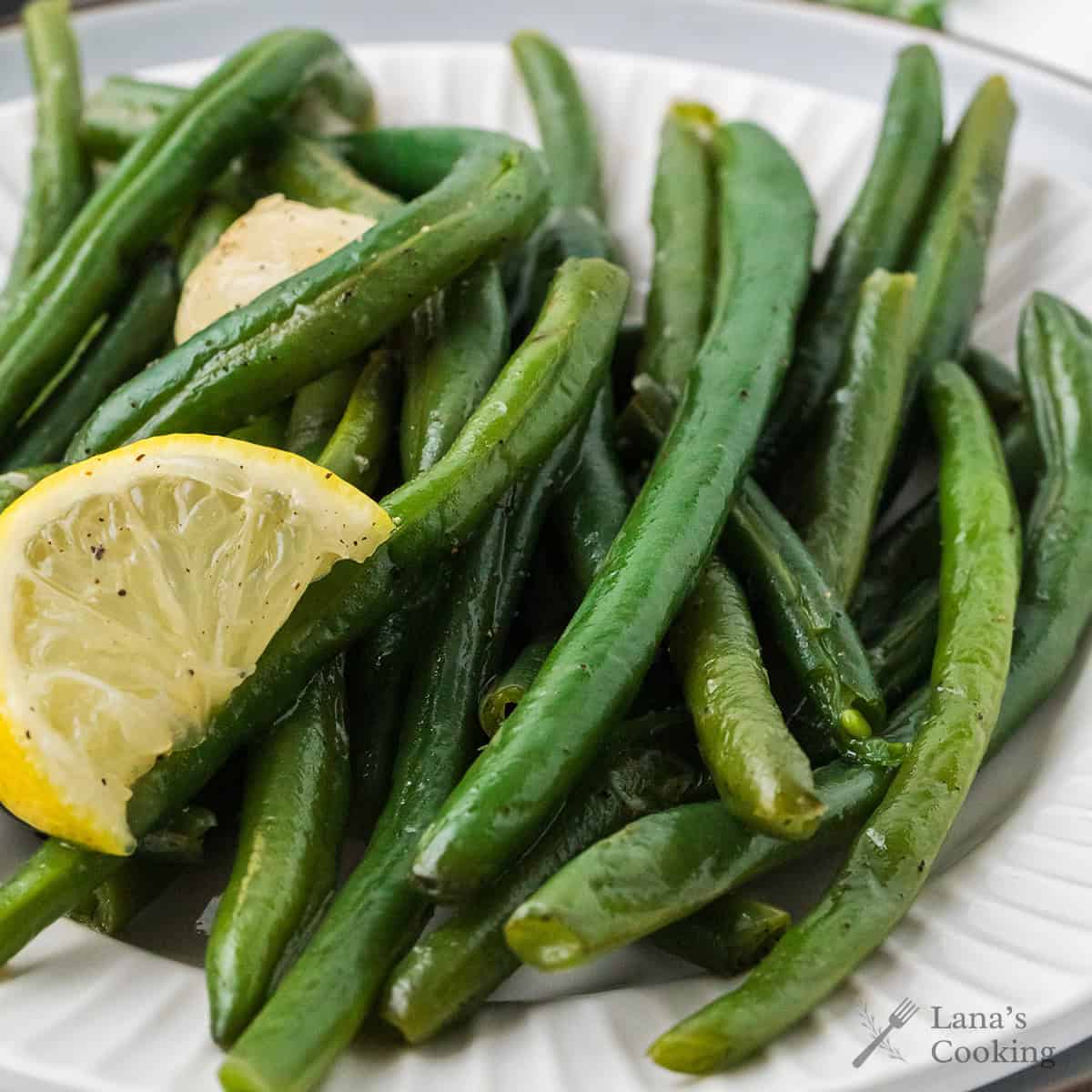 Green beans garnished with a lemon wedge and garlic on a white plate.