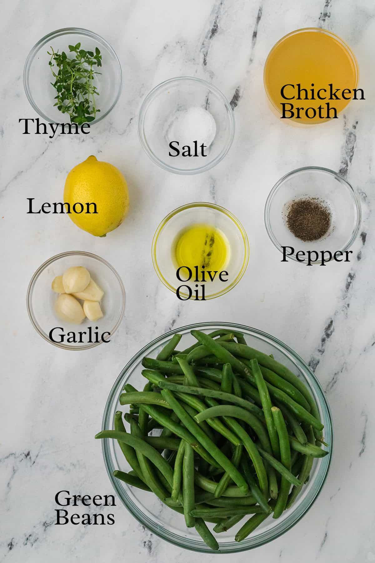 Bowls of green beans, garlic, thyme, lemon, olive oil, chicken broth, salt, and pepper on a marble counter.