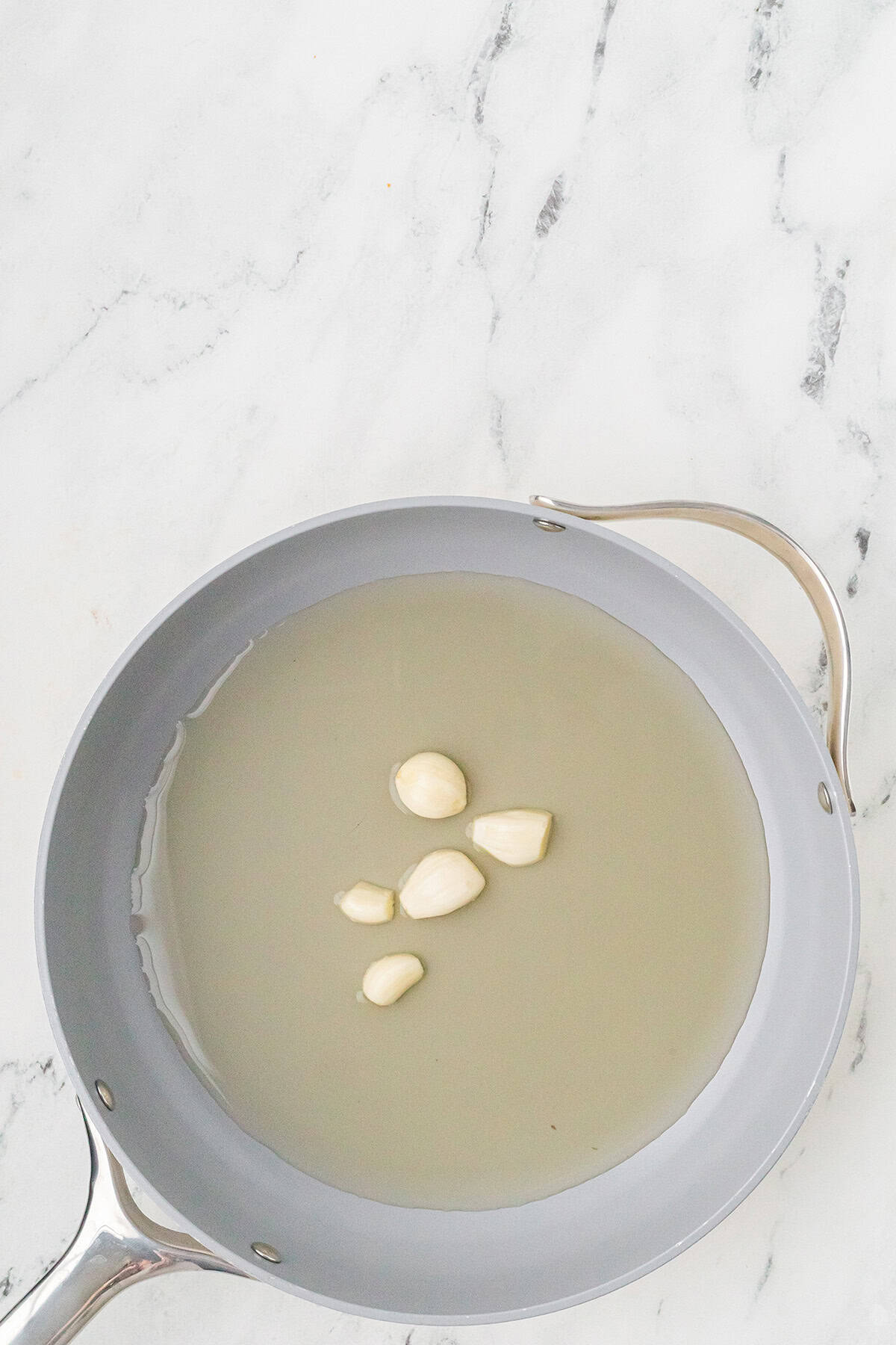 A frying pan with chicken broth and several garlic cloves on a white marble surface.