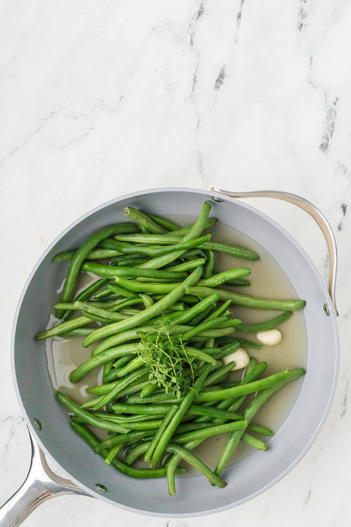 Green beans, garlic cloves, and fresh herbs cooking in broth in a silver pan on a marble surface.