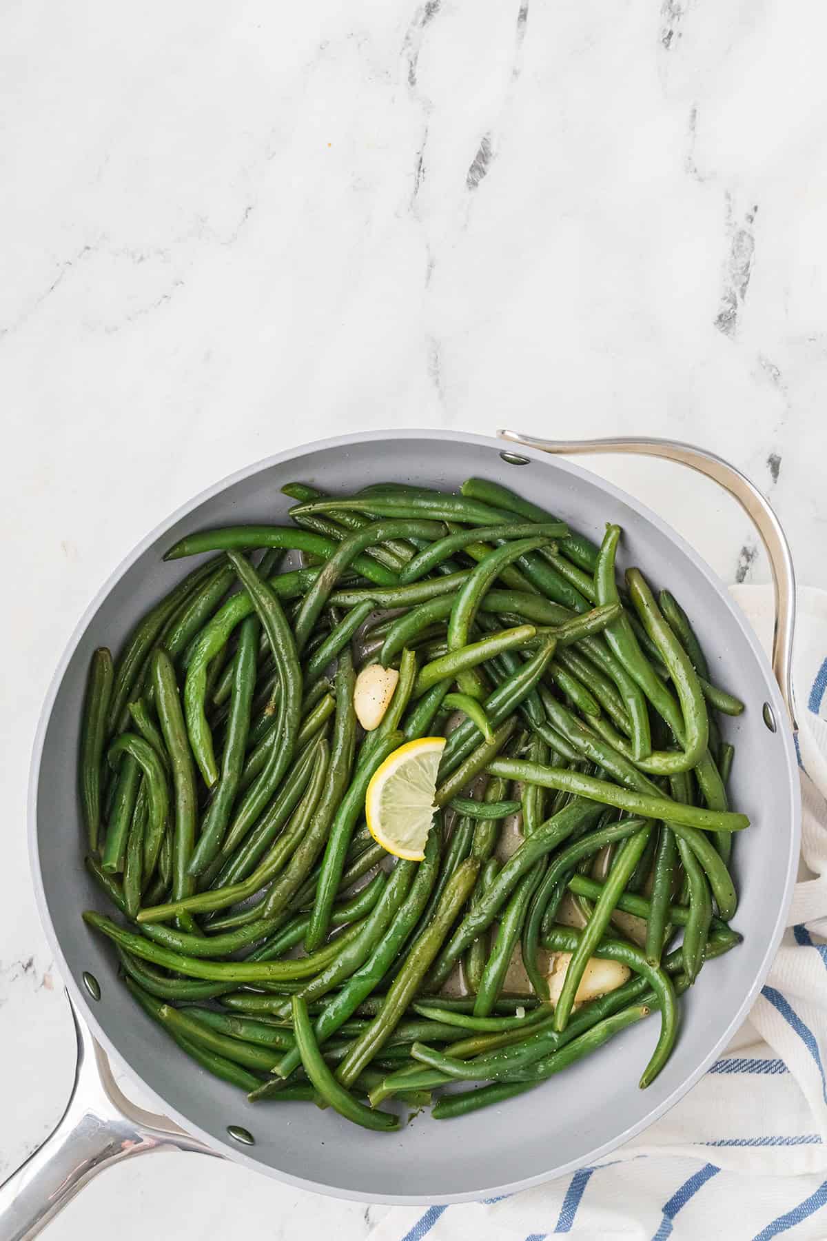 Green beans braised with garlic and a lemon slice in a skillet on a marble countertop.