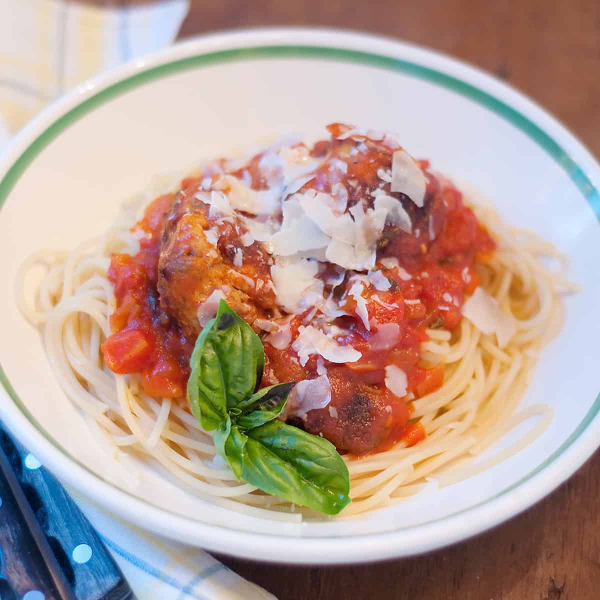 A plate of spaghetti topped with tomato sauce, meatballs, shaved parmesan cheese, and a sprig of basil, served in a white bowl with a green rim on a wooden table.
