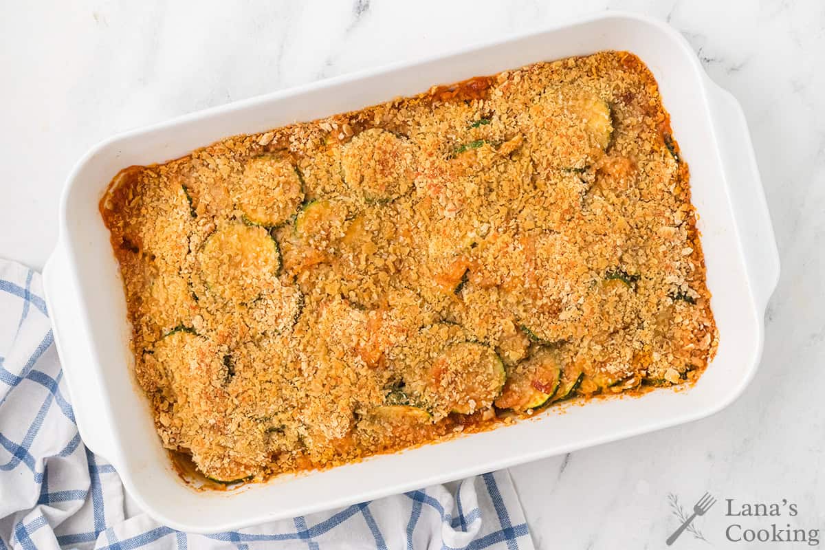 Baked zucchini casserole with golden cracker topping in a white dish on a marble surface with a towel.