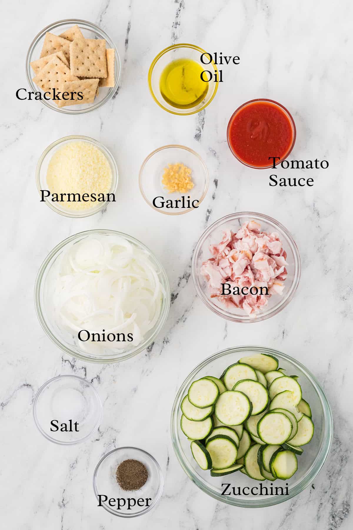Bowls of crackers, olive oil, tomato sauce, parmesan, garlic, bacon, onions, zucchini, salt, and pepper on a counter.