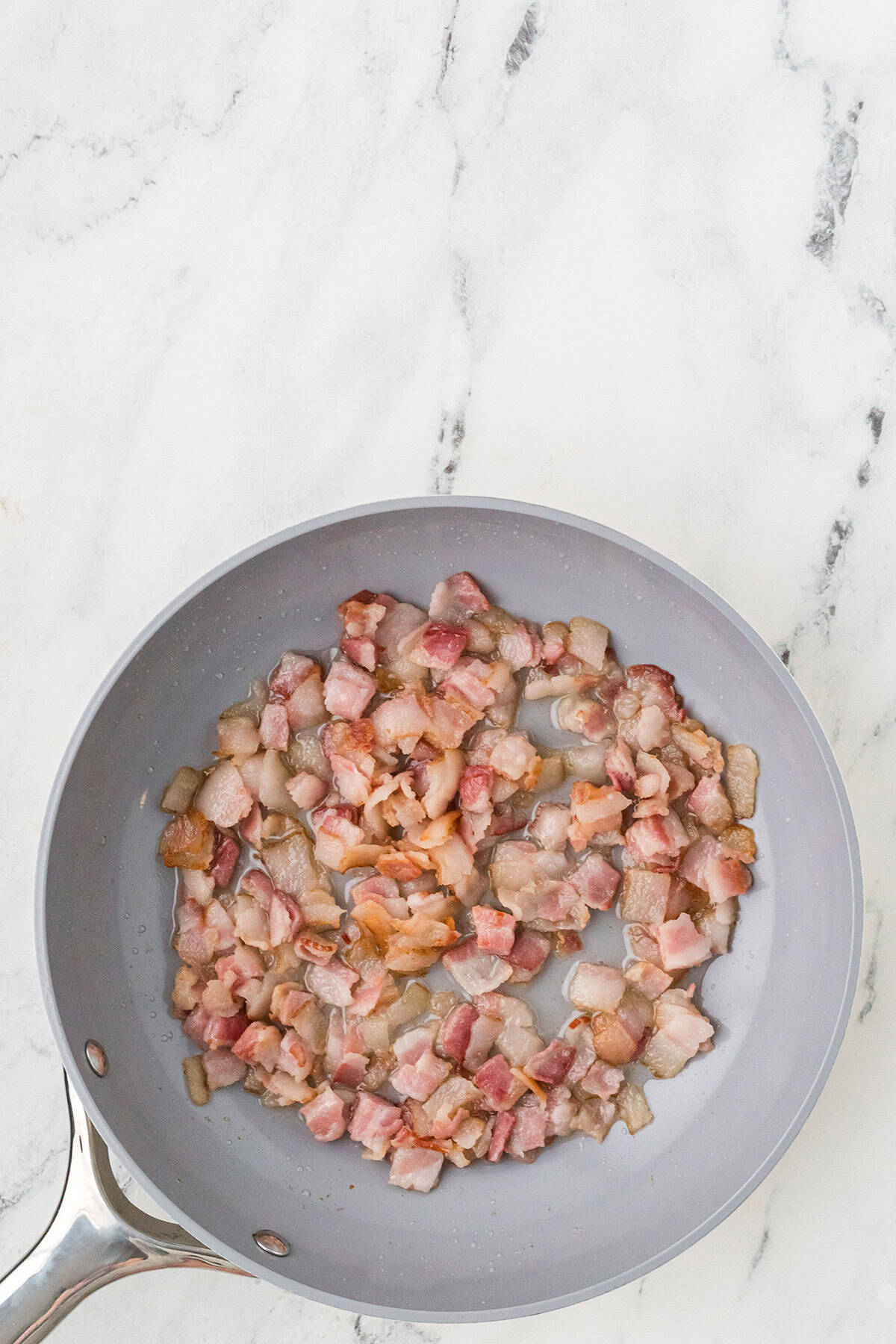 Chopped bacon pieces cooking in a gray frying pan on a white marble countertop.