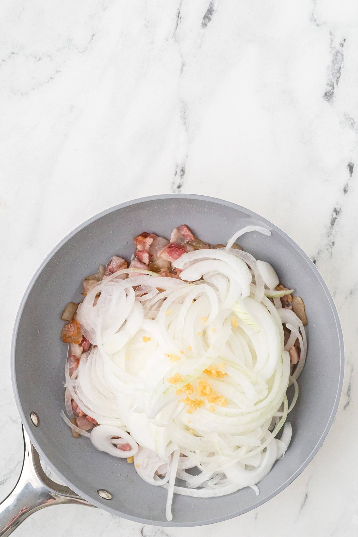 Sliced onions, minced garlic, and sour cream on top of cooked bacon in a gray skillet on a marble surface.