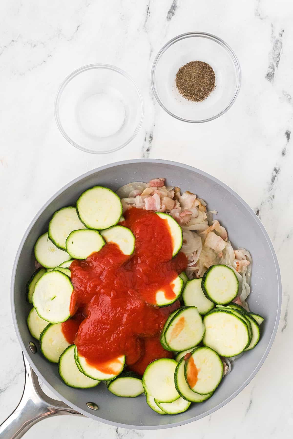 A pan with zucchini, tomato sauce, onions, and bacon, with bowls of salt and pepper on a marble surface.