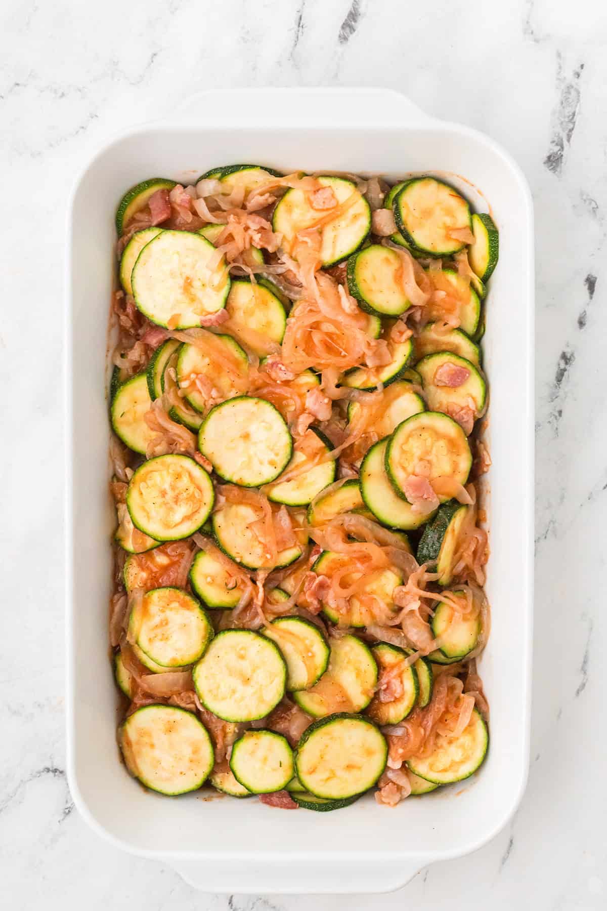 Sliced zucchini with tomatoes and onions in a white baking dish on a marble surface.
