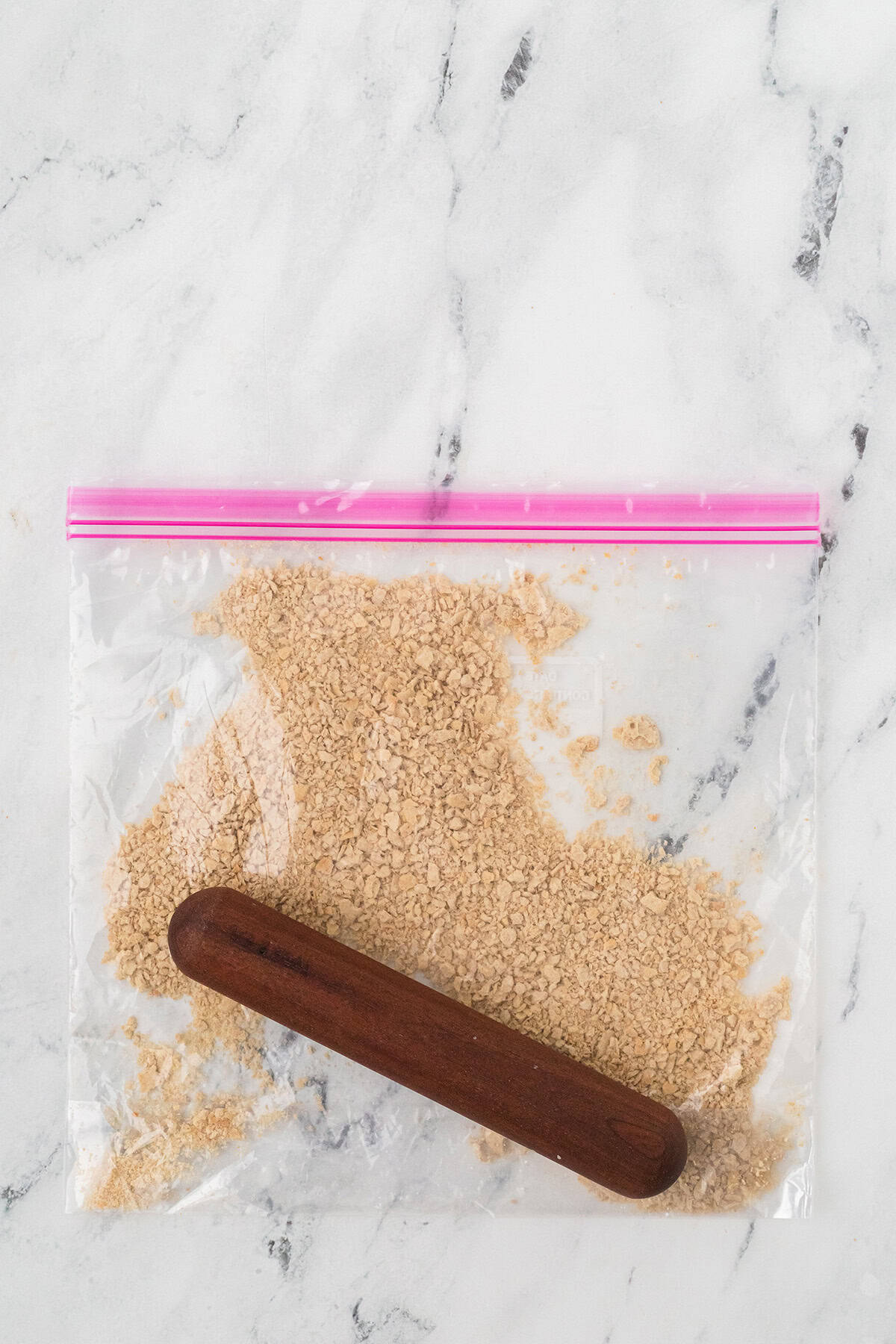 A wooden rolling pin crushing crackers inside a clear zip-top plastic bag on a marble surface.