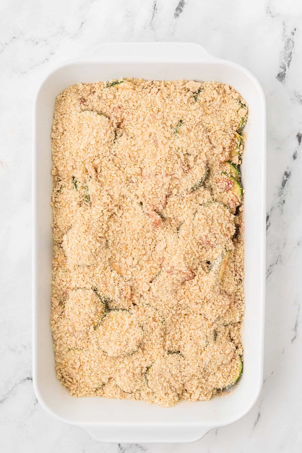 A white baking dish filled with a cracker crumb-topped casserole, ready to be baked, on a marble surface.