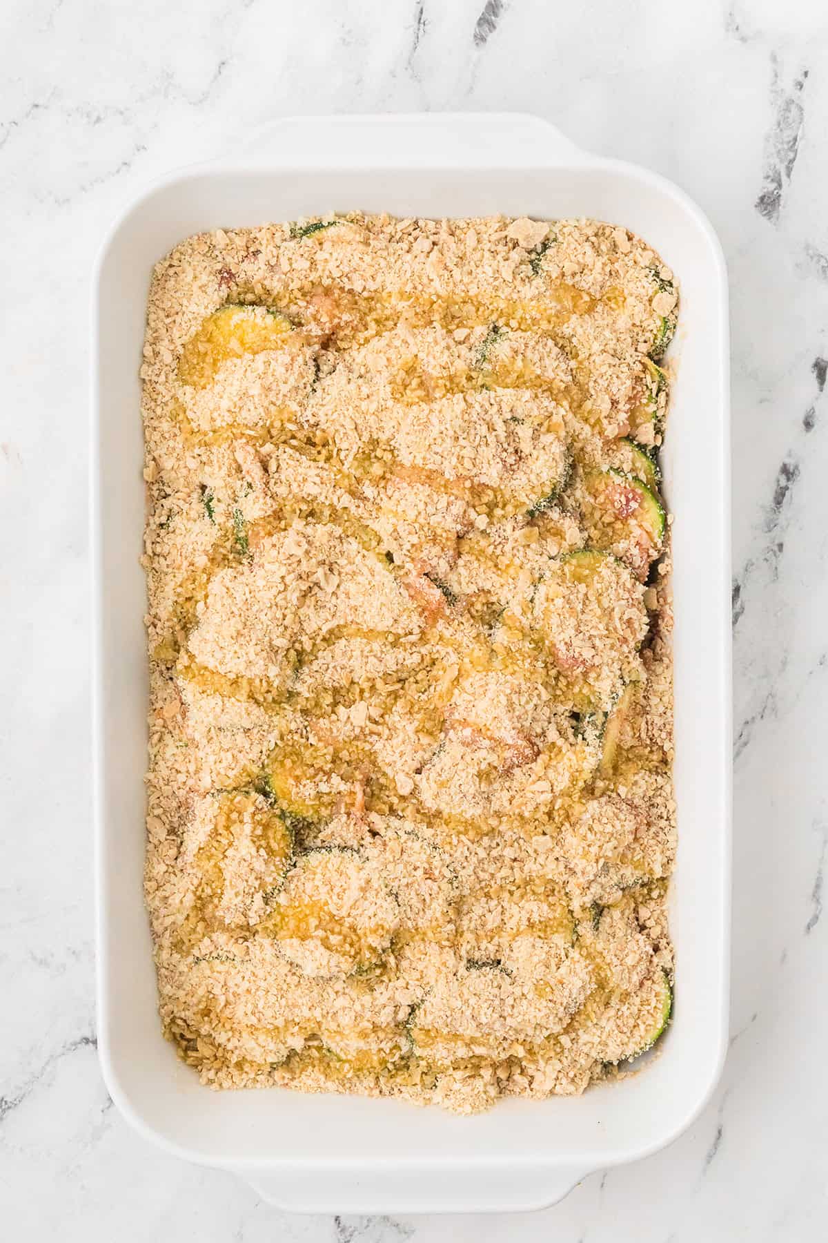 Rectangular white baking dish with a cracker topping drizzled with olive oil over a layered casserole on a marble surface.