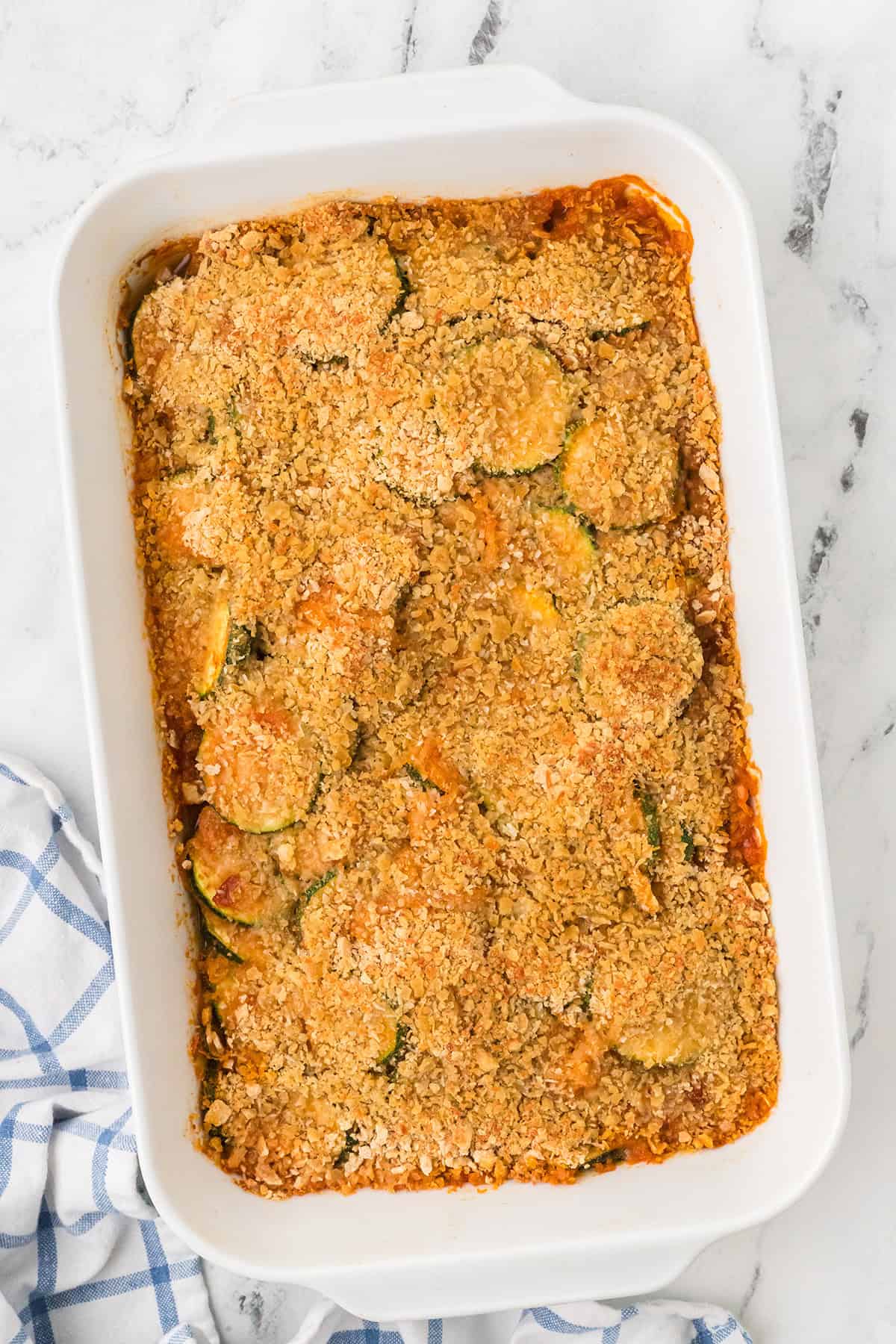 Baked casserole with a golden cracker topping in a white dish on a marble surface with a blue checked towel.