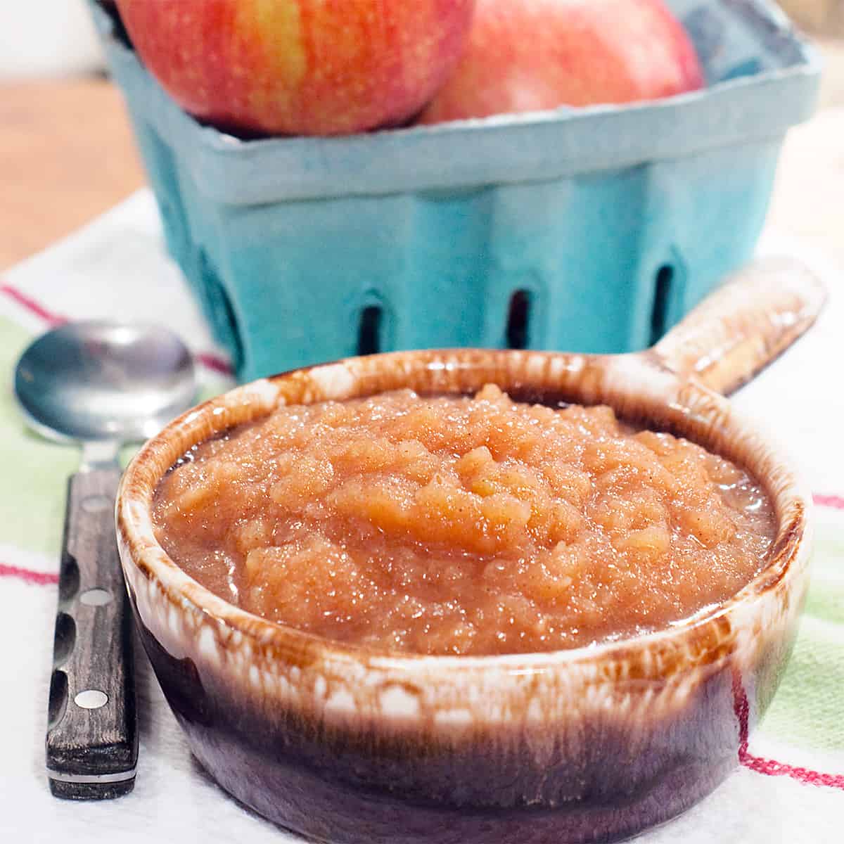 Bowl of applesauce with a basket of fresh apples on a kitchen towel.