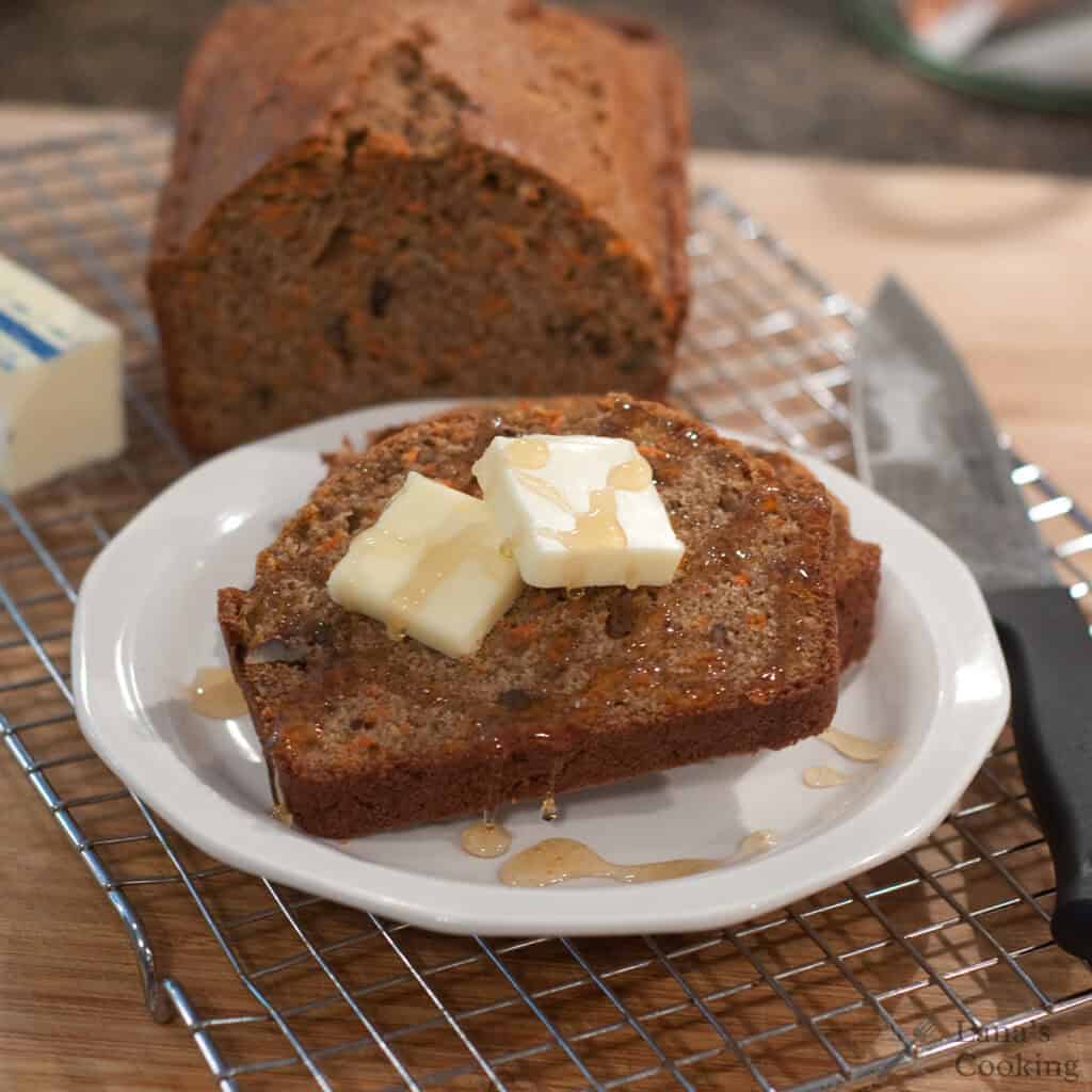 A slice of carrot pecan quick bread topped with butter and honey on a white plate, with a whole loaf in the background.