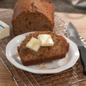 A slice of carrot pecan quick bread topped with butter and honey on a white plate, with a whole loaf in the background.