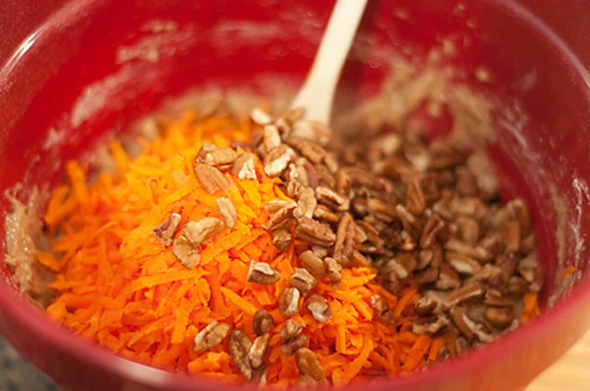 Shredded carrots and pecans in a red mixing bowl with a wooden spoon.