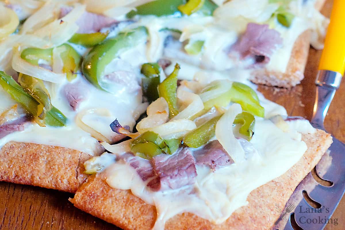 A close-up of a sliced flatbread topped with melted cheese, green bell peppers, onions, and pieces of cooked meat on a wooden surface.