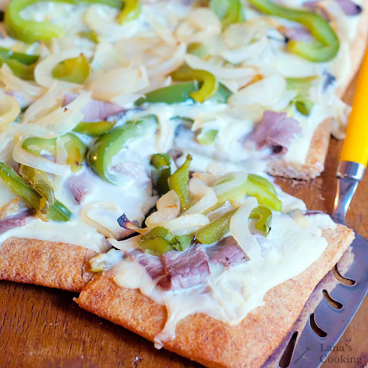 A rectangular pizza with a wheat crust, topped with melted cheese, green bell peppers, onions, and slices of meat, with one piece being lifted by a spatula.