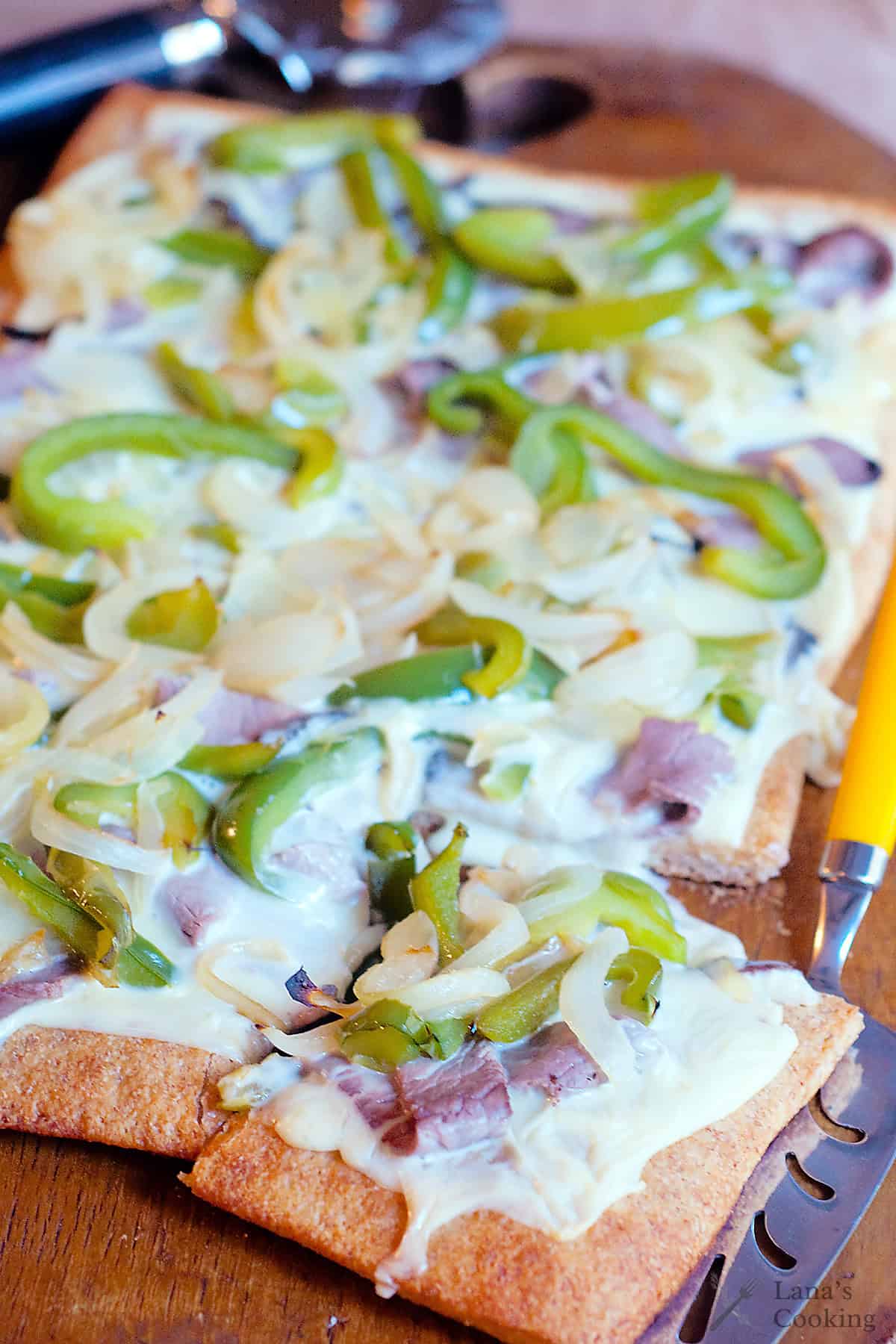 Rectangular flatbread pizza topped with melted cheese, green bell peppers, onions, and pieces of meat, served on a wooden board with a yellow-handled knife.