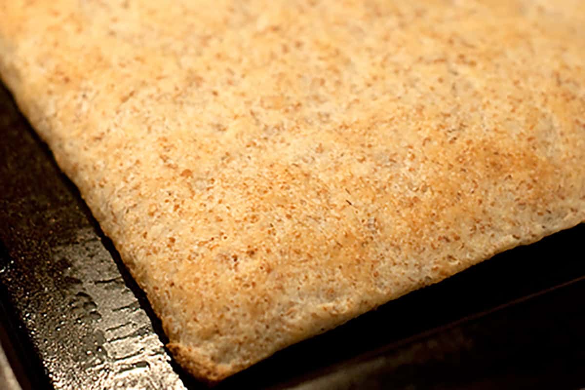 A close-up of a rectangular, golden-brown baked pizza crust on a dark metal baking tray.