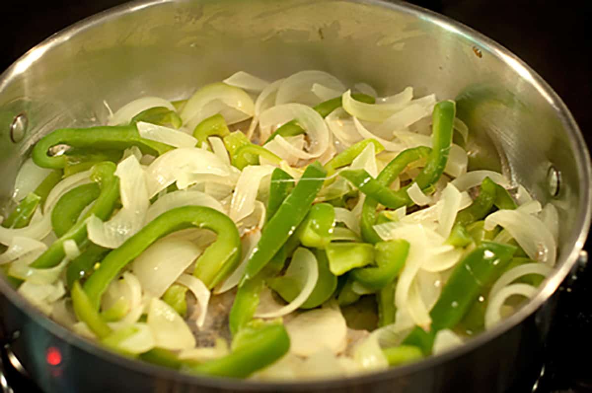 Sliced green bell peppers and onions are being sautéed in a stainless steel pan.