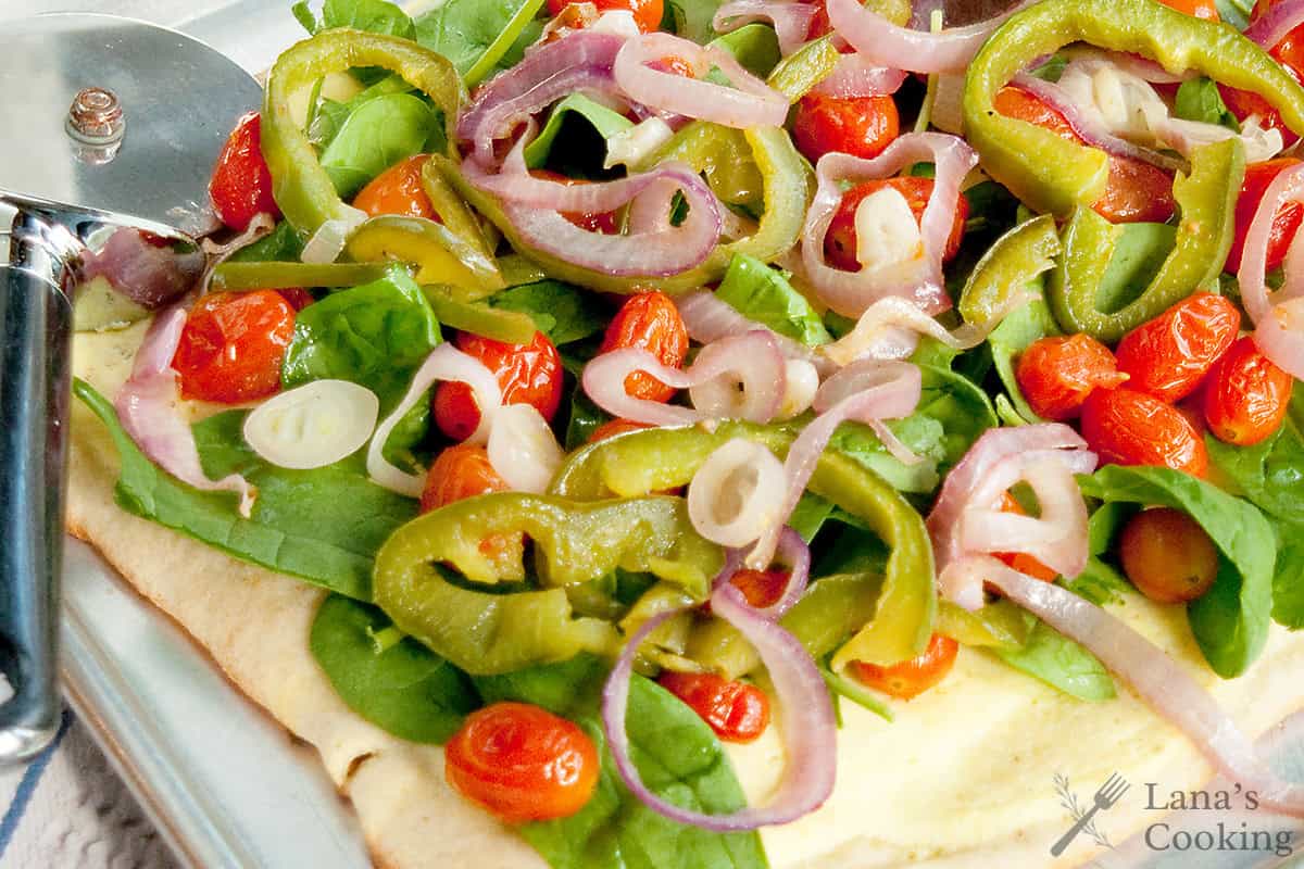 A close-up of a vegetable pizza topped with spinach, cherry tomatoes, green peppers, and red onions next to a pizza cutter on a metal tray.