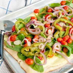 A rectangular flatbread topped with spinach, cherry tomatoes, sliced red onions, and green peppers on a baking sheet with a pizza cutter beside it.