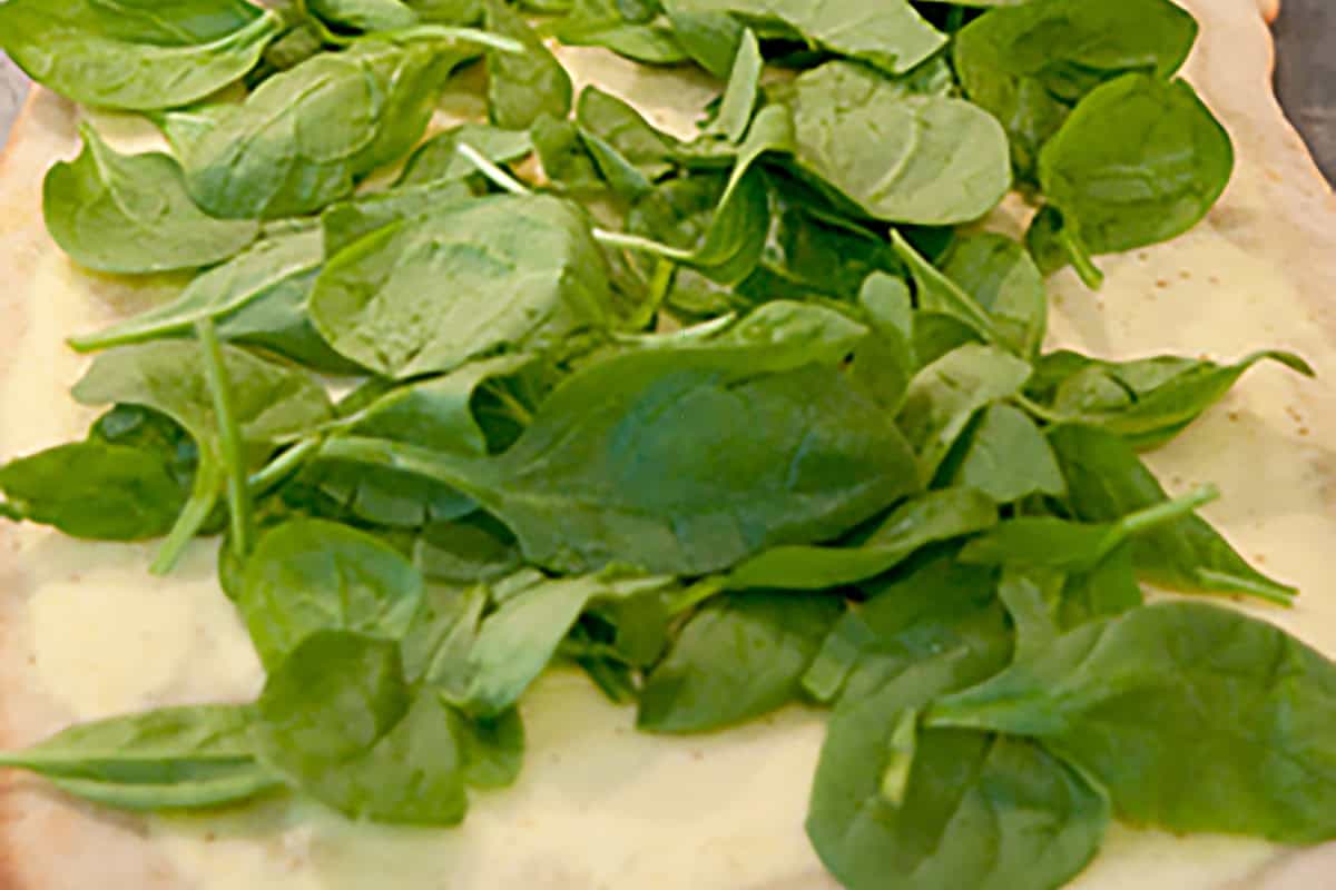 A rectangular pizza crust on a baking sheet topped with ricotta and fresh spinach leaves.