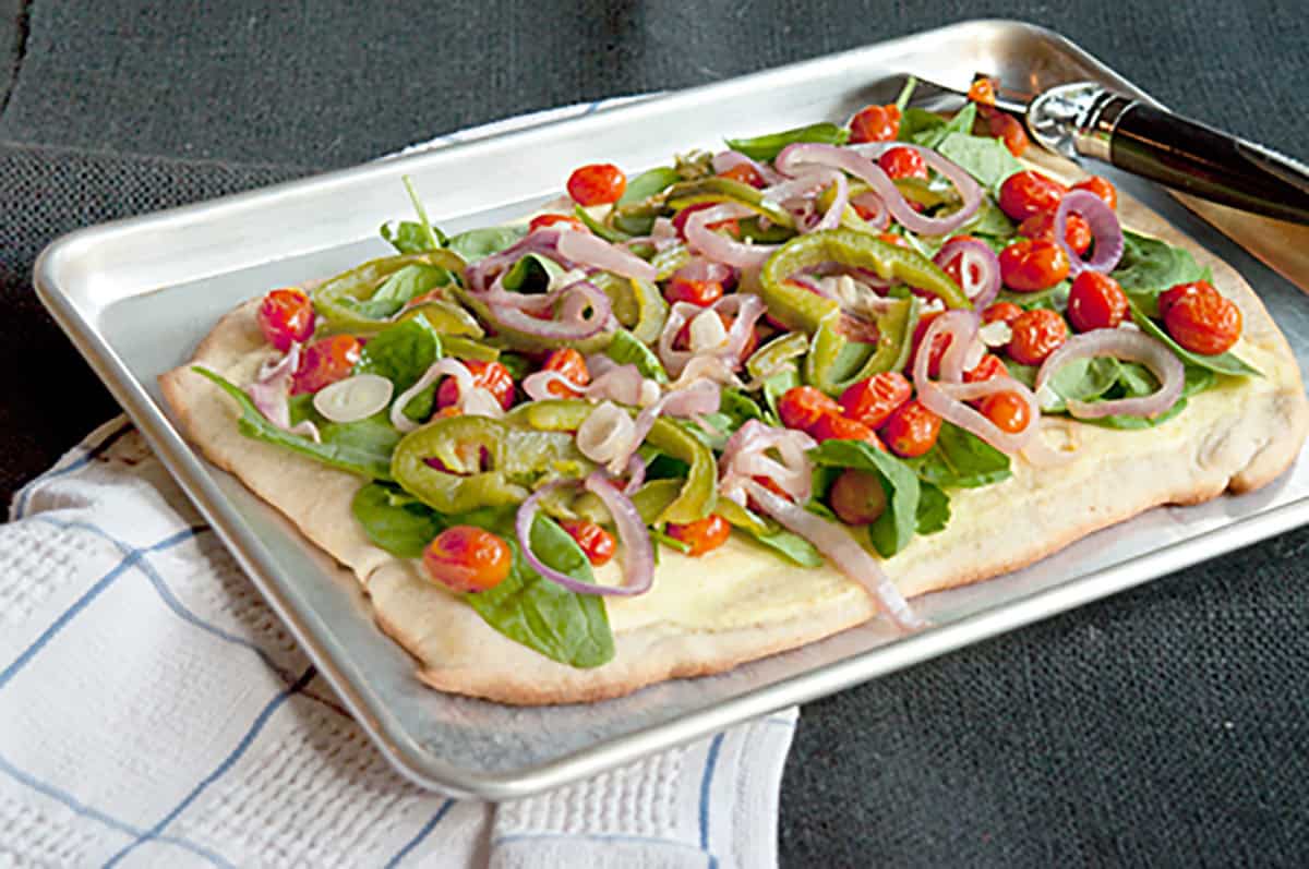 Rectangular pizza topped with spinach, cherry tomatoes, green peppers, and sliced red onions on a baking tray with a towel and serving utensil beside it.