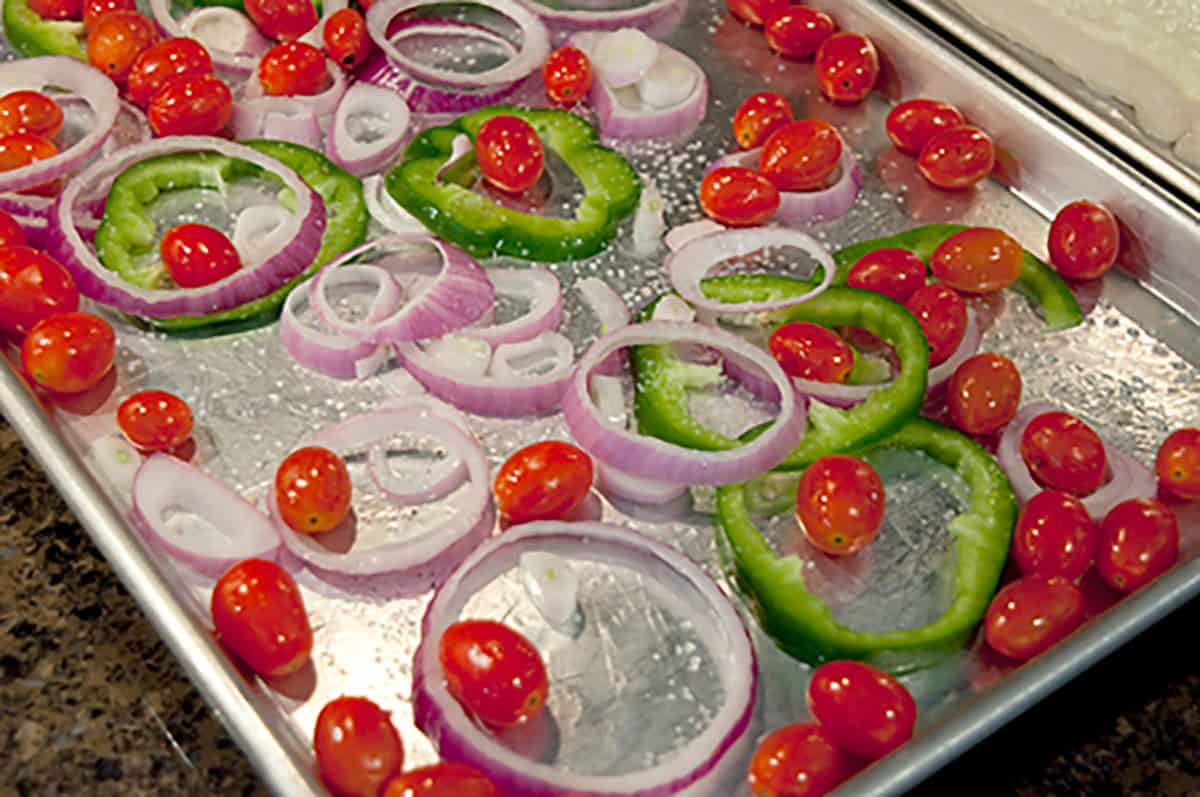 Sliced red onions, green bell peppers, and cherry tomatoes arranged on a baking sheet, ready for roasting.